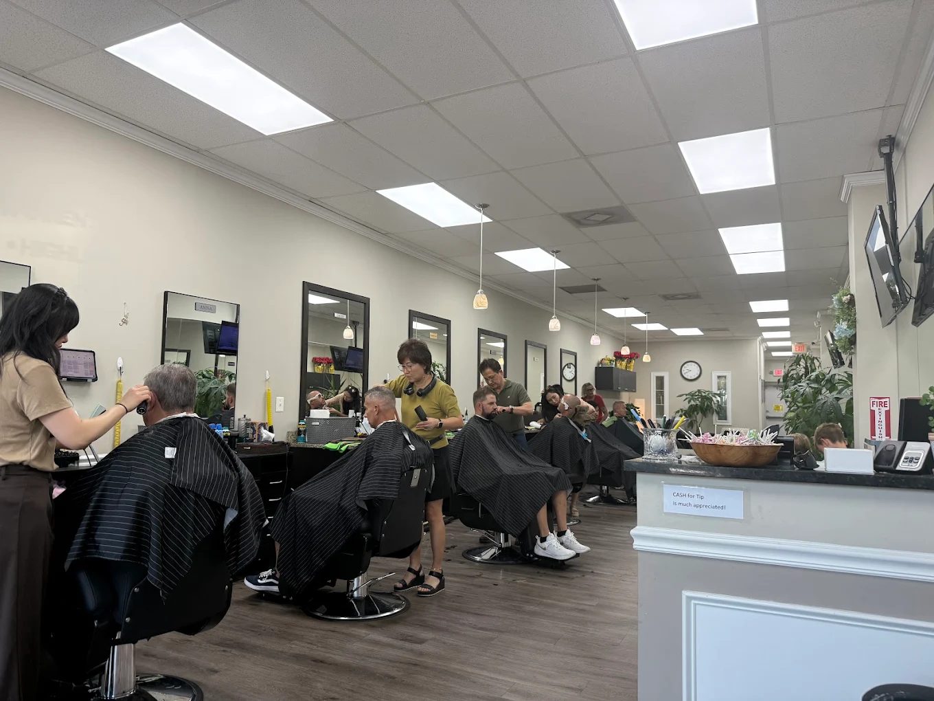 Barbers working down the length of the shop with most chairs full, mirrors and televisions along the wall.