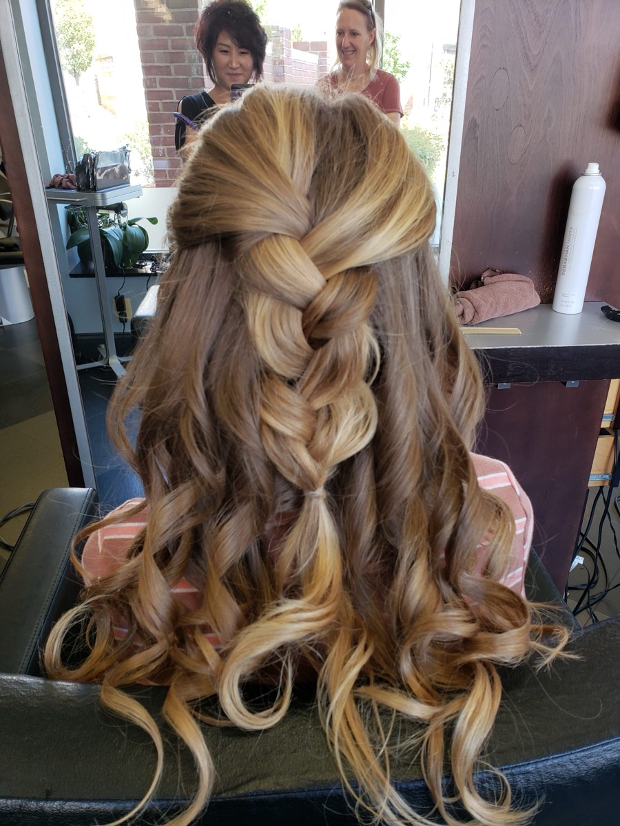A long braided half-up style with loose curls, finished in the shop with the storefront windows and brick behind.